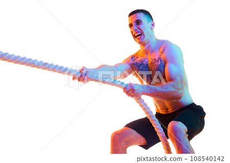 Muscular shirtless young man with strong body and hands pulling rope, training against white studio background in neon light Muscular shirtless young man with strong body and hands pulling rope, training against white studio background in neon light 108540142
