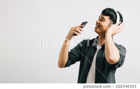 Happy man sings with delight while using Bluetooth headphones for karaoke on his smartphone. Studio shot isolated on white, showcasing his passion for music and technology. Happy man sings with delight while using Bluetooth headphones for karaoke on his smartphone. Studio shot isolated on white, showcasing his passion for music and technology. 108540391