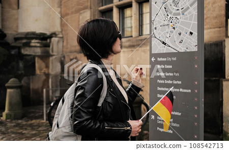 Attractive Female Tourist Walks With Germany Flag In Historical Center Of Dresden, Checking Touristic Route On City Map 108542713