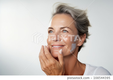 Close-up of middle-aged Caucasian woman touching her face to apply moisturizer. Smiling face of adult grey-haired lady with daily cream, facial cosmetics. Skin care. Grey background, copy space. Close-up of middle-aged Caucasian woman touching her face to apply moisturizer. Smiling face of adult grey-haired lady with daily cream, facial cosmetics. Skin care. Grey background, copy space. 108544395