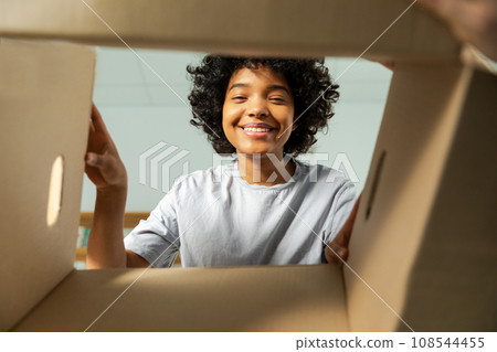 Inside of cardbox bottom view. African girl unpacking delivery looking in box. Woman opening carton box. Female getting parcel looking at delivered goods items. Satisfied client positive feedback Inside of cardbox bottom view. African girl unpacking delivery looking in box. Woman opening carton box. Female getting parcel looking at delivered goods items. Satisfied client positive feedback 108544455