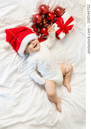 Christmas toddler in Santa hat lying on bed with gift box and festive baubles top view Christmas toddler in Santa hat lying on bed with gift box and festive baubles top view 108544702