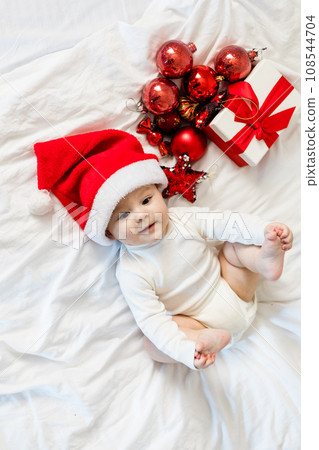 Christmas toddler in Santa hat sitting on bed with gift box and festive baubles Christmas toddler in Santa hat sitting on bed with gift box and festive baubles 108544704