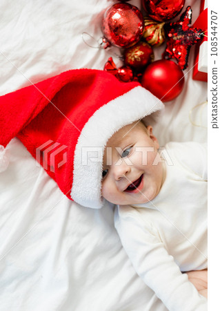 Christmas toddler in Santa hat lying on bed with gift box and festive baubles top view 108544707