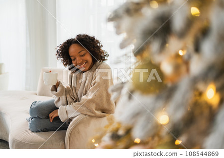 Merry Christmas. African American woman with cup of hot drink coffee tea sitting near Christmas tree at home. Girl in living room with Christmas tree. Christmas hygge holidays at home Merry Christmas. African American woman with cup of hot drink coffee tea sitting near Christmas tree at home. Girl in living room with Christmas tree. Christmas hygge holidays at home 108544869