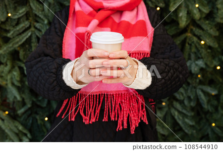 A young woman in a bright pink scarf holds a disposable glass of coffee in her hand while walking against the background of a wall of a Christmas tree with a garland. 108544910