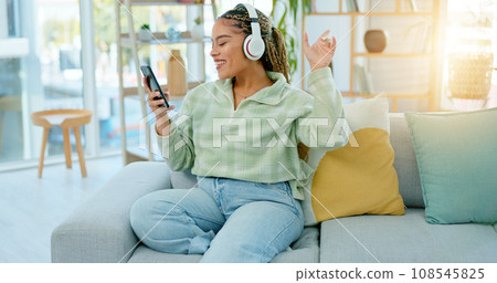 Phone, sofa and happy woman on music headphones in home living room for dancing. Mobile, radio and African person listening to podcast, audio and sound for freedom, celebration and streaming online Phone, sofa and happy woman on music headphones in home living room for dancing. Mobile, radio and African person listening to podcast, audio and sound for freedom, celebration and streaming online 108545825