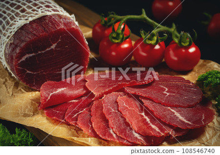 Whole and sliced bresaola on paper on a cutting board with tomatoes Whole and sliced bresaola on paper on a cutting board with tomatoes 108546740