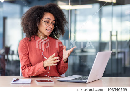 Female boss with wired headphones headset talking remotely with colleagues, using laptop for video call, businesswoman in red shirt smiling satisfied inside office at workplace. 108546769