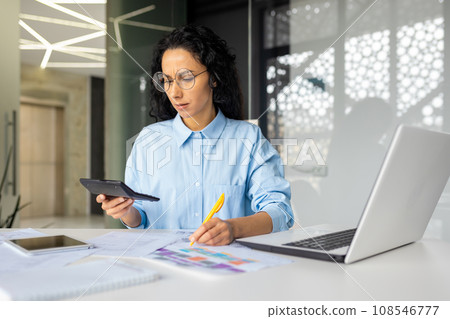 Upset frustrated financial woman checking paper reports, businesswoman behind paperwork inside office, using calculator and laptop to check and analyze report. Upset frustrated financial woman checking paper reports, businesswoman behind paperwork inside office, using calculator and laptop to check and analyze report. 108546777