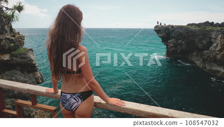 Long hair brunette in bikini standing on a balcony with awesome views of ocean rock cliff. Tourist beautiful woman relaxes on seashore cliff. Travel, tourism, holiday. Back view 108547032