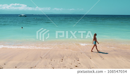 Young woman taking a peaceful stroll along a serene tropical beach. Girl walking white sand, turquoise sea water in background. Amazing natural summer scenery. Travel, tourism, holiday. 108547034