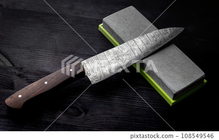 Big Kitchen Knife Rests On Sharpening Stone On Kitchen Table Against Black Background 108549546