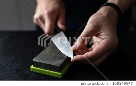Man Uses Natural Sharpening Whetstone To Sharpen Big Kitchen Knife Against Black Background Man Uses Natural Sharpening Whetstone To Sharpen Big Kitchen Knife Against Black Background 108549547