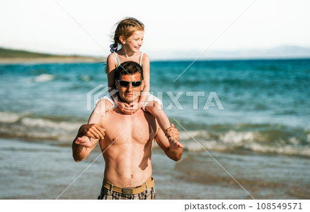 Father holding daughter at shoulders at sea coast portraits. Family dad and little pretty child kid at beach at summer together Father holding daughter at shoulders at sea coast portraits. Family dad and little pretty child kid at beach at summer together 108549571
