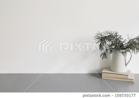 Elegant Christmas interior, home office. Ceramic vase, jug with green pine tree branches on old books. Winter home decor. Empty white wall background mockup. Grey table, desk. No people. 108550177