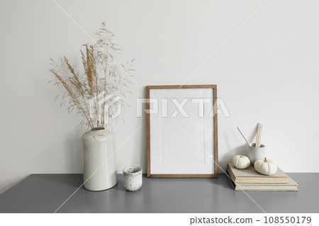 Elegant interior, home office. Blank wood picture frame mockup. Cup of coffee, pencils and little pumpkins. Vase with dry grass. Autumn boho home decor. White wall background. Grey table, desk. Elegant interior, home office. Blank wood picture frame mockup. Cup of coffee, pencils and little pumpkins. Vase with dry grass. Autumn boho home decor. White wall background. Grey table, desk. 108550179