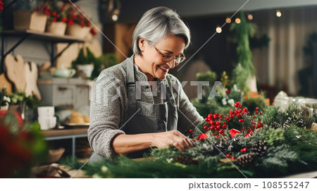 An older woman florist makes and decorates a Christmas wreath from fir branches with red berries and holly branches 108555247