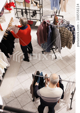 Senior people browsing for clothes on hangers and looking for perfect formal outfit to attend christmas dinner festivity. Elderly couple doing shopping spree at mall during sales season. Senior people browsing for clothes on hangers and looking for perfect formal outfit to attend christmas dinner festivity. Elderly couple doing shopping spree at mall during sales season. 108558114