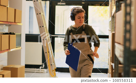 Depot manager having fun in warehouse, reviewing inventory list and listening to music while she works in storage room. Young adult checking retail order shipment, supply chain. Handheld shot. 108558311