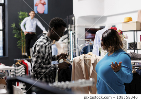 Stylish client shopping for fashion collection, looking at shirt fabric in modern boutique. African american couple buying fashionable clothes, checking hangers with formal wear in clothing store Stylish client shopping for fashion collection, looking at shirt fabric in modern boutique. African american couple buying fashionable clothes, checking hangers with formal wear in clothing store 108558346