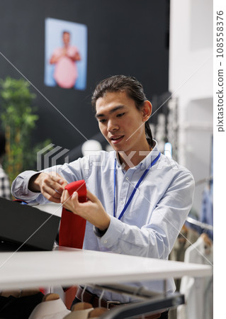 Asian man looking at fashionable accessories, arranging belt on store shelf in modern boutique. Stylish employee working in shopping centre, working with hangers full with sales clothes Asian man looking at fashionable accessories, arranging belt on store shelf in modern boutique. Stylish employee working in shopping centre, working with hangers full with sales clothes 108558376
