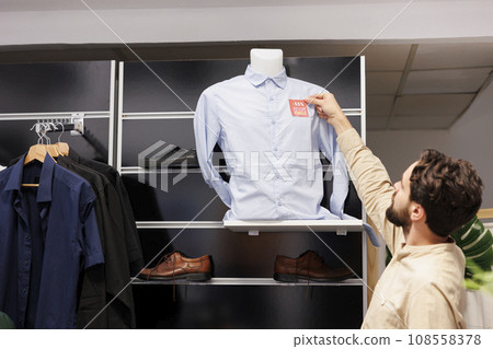 Young man reading red sale price tag while shopping for clothes on Black Friday in fashion mall, male customer standing in clothing store clearance section looking at discounted shirt Young man reading red sale price tag while shopping for clothes on Black Friday in fashion mall, male customer standing in clothing store clearance section looking at discounted shirt 108558378