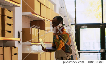 African american man inspecting products and talking about distribution with headquarters, cargo stock. Male supervisor using landline phone in storage room, quality control verification. African american man inspecting products and talking about distribution with headquarters, cargo stock. Male supervisor using landline phone in storage room, quality control verification. 108558380