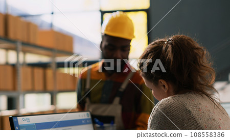 Manager reviewing distribution papers and discussing order delivery with young employee, boxes of products in storage room. Diverse team of depot workers preparing stock inventory. 108558386