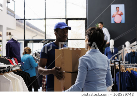 African american courier holding cardboard boxes, discussing shipping detalis with woman manager in shopping centre. Man in delivery uniform working with fashion store. Stylish boutique concept African american courier holding cardboard boxes, discussing shipping detalis with woman manager in shopping centre. Man in delivery uniform working with fashion store. Stylish boutique concept 108559748