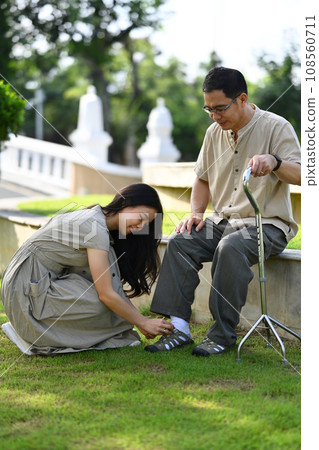 Caring young adult daughter helping her father tying shoelaces while relaxing in the public park Caring young adult daughter helping her father tying shoelaces while relaxing in the public park 108560711