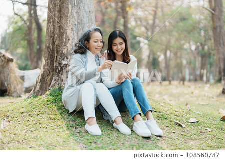 Grown daughter with aging mother expressing love and looking at memory notebook under tree in public park. 108560787