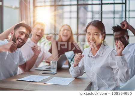 group of different business people celebrating victory while sitting at table in modern office group of different business people celebrating victory while sitting at table in modern office 108564990