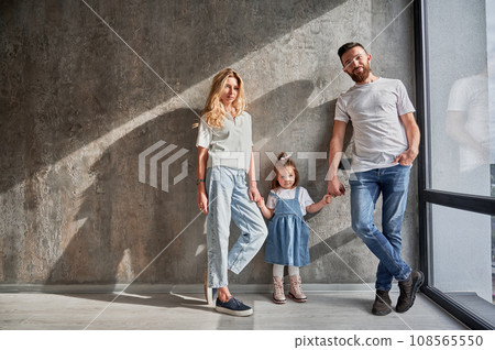 Portrait of happy parents and child standing by the window against the wall in apartment. Man and woman holding hands of adorable daughter in new home with sunlight shadow on the wall. 108565550