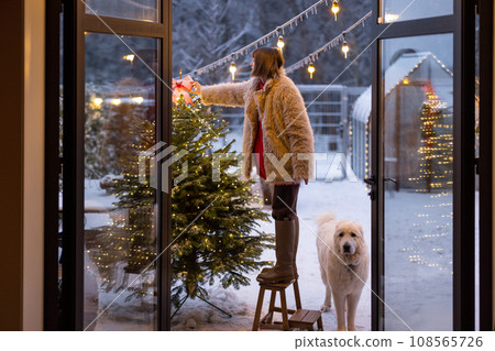 Woman decorates Christmas tree on house terrace outdoors Woman decorates Christmas tree on house terrace outdoors 108565726