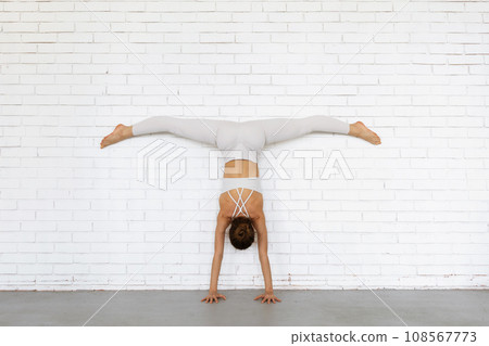 Young woman trainer practicing yoga doing Adho Mukha Vrikshasana exercise with Samakonasana, handstand with transverse splits with legs against the wall, inverted asana, exercising in white clothes Young woman trainer practicing yoga doing Adho Mukha Vrikshasana exercise with Samakonasana, handstand with transverse splits with legs against the wall, inverted asana, exercising in white clothes 108567773