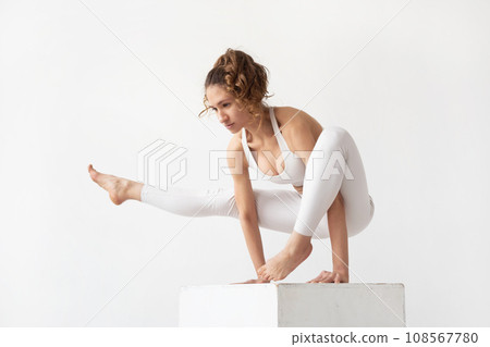Young woman leading a healthy lifestyle and practicing yoga, performs the exercise eka pada bhujapidasana, handstand on a white cube, trains in white sportswear on a light background Young woman leading a healthy lifestyle and practicing yoga, performs the exercise eka pada bhujapidasana, handstand on a white cube, trains in white sportswear on a light background 108567780