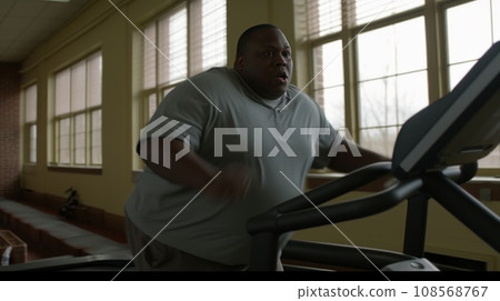 Overweight man walk on treadmill in sports club with panoramic windows overlooking sunny city Overweight man walk on treadmill in sports club with panoramic windows overlooking sunny city 108568767