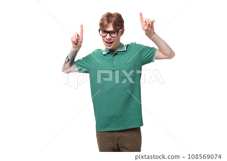young surprised caucasian male student with red hair and tattoo reports news and points up with 108569074