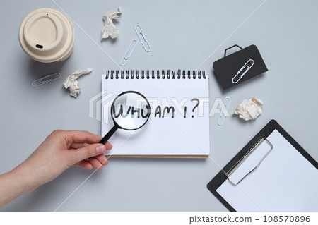 Notepad with the text Who am I?, magnifying glass in hand, papers and cup on gray background, top view Notepad with the text Who am I?, magnifying glass in hand, papers and cup on gray background, top view 108570896
