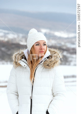 Portrait of serious young woman in white jacket and knitted hat in snowy winter day. Winter walk. Vertical frame. 108572797