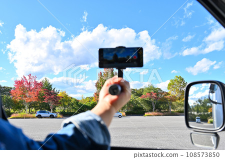 A woman taking pictures with a smartphone from inside a car, live streaming autumn leaves 108573850
