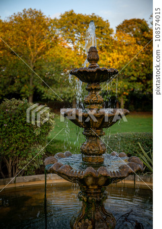 Fountain in a park with the setting sun illuminating the background trees. Portrait view. 108574015