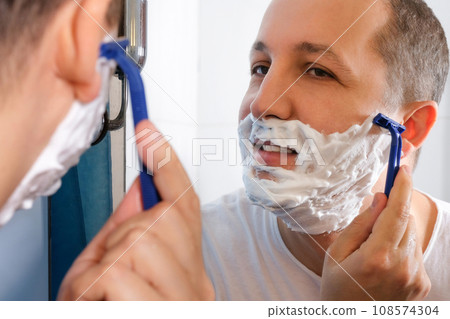 Young man with foam on his face shaves in front of a mirror. Young man with foam on his face shaves in front of a mirror. 108574304