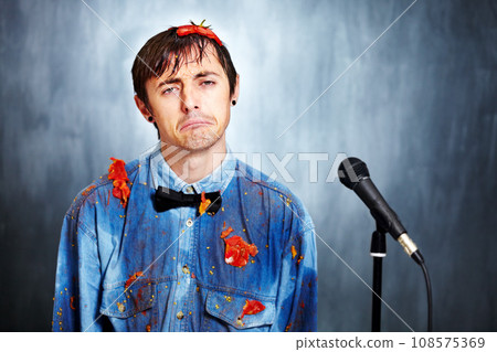 Portrait, sad and a man comic in tomato with a microphone after a bad performance at a comedy club. Depression, fail or boo with an unhappy young comedian in studio on a gray background for a show Portrait, sad and a man comic in tomato with a microphone after a bad performance at a comedy club. Depression, fail or boo with an unhappy young comedian in studio on a gray background for a show 108575369
