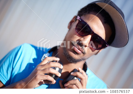 Portrait, fashion and music with a man in sunglasses outdoor on a blurred background for audio streaming. Face, headphones and hip hop with a confident young person in street wear for urban style 108576537