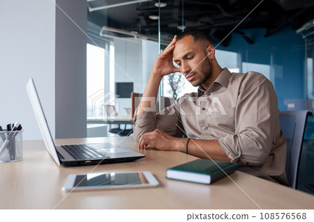 Upset tired young african american man sitting at desk with laptop in office, tired of work holding hand on head, suffering from headache, bored in office. Upset tired young african american man sitting at desk with laptop in office, tired of work holding hand on head, suffering from headache, bored in office. 108576568