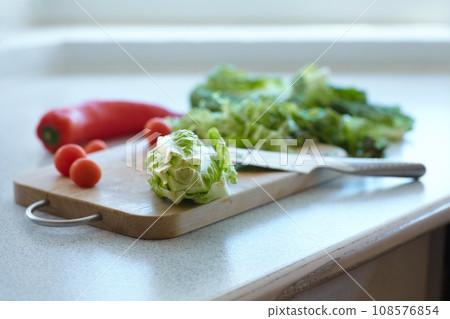 Chopping board, vegetables and knife on table in kitchen cooking organic food. Cutting wood, blade and plants on counter, lettuce and tomatoes, pepper and nutrition for healthy diet or salad in home Chopping board, vegetables and knife on table in kitchen cooking organic food. Cutting wood, blade and plants on counter, lettuce and tomatoes, pepper and nutrition for healthy diet or salad in home 108576854