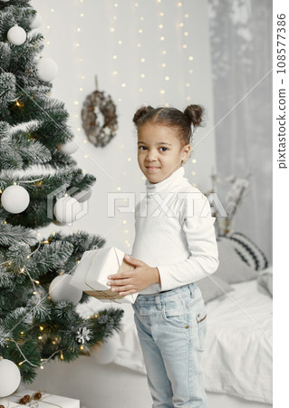 Child in a white sweater. Daughter standing near Christmas tree. 108577386
