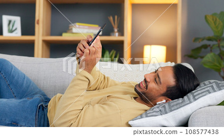 Man, phone and earphones to relax on sofa, texting or reading with smile, contact or music in home living room. Guy, chat and smartphone for audio streaming subscription, video or comic meme on couch 108577438
